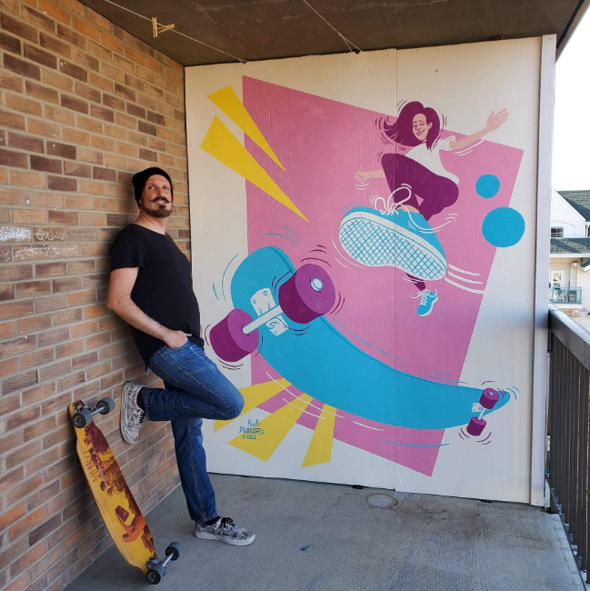Photo of artist Rob Milton beside his mural of a girl skateboarding with 90's graphics and colours.